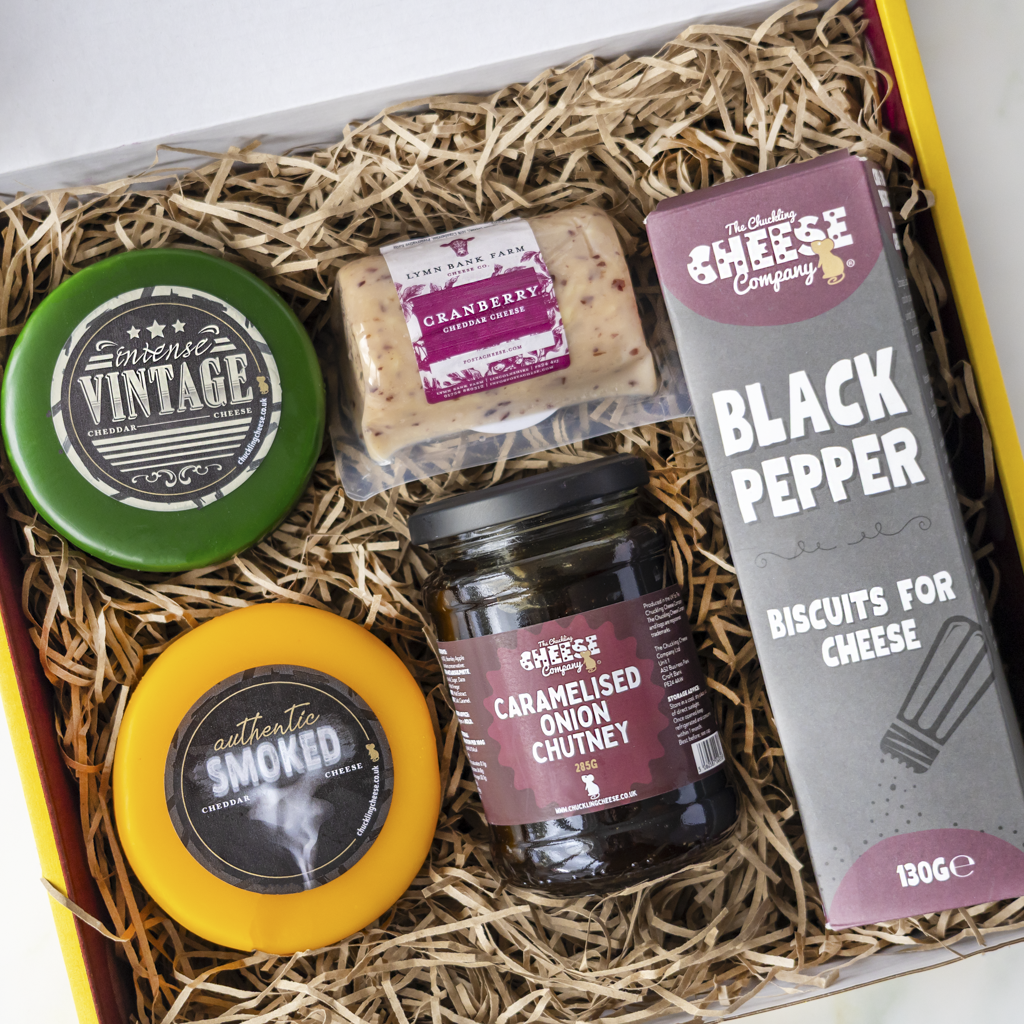 A gourmet cheese and biscuit gift box from The Chuckling Cheese Company, filled with shredded paper packaging. The box contains a green 'Vintage' cheese wheel, a yellow 'Smoked' cheese wheel, a jar of caramelised onion chutney, a package of cranberry cheese crisp, and a box of black pepper biscuits.