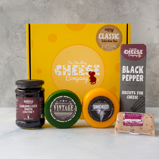A Chuckling Cheese Company gift box set arranged on a white surface against a grey textured wall. The set includes a yellow branded box, a jar of caramelised onion chutney, three round cheese wheels in green, yellow, and orange packaging, and a package of black pepper biscuits.