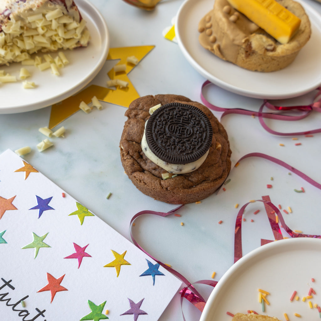 A decorated chocolate chip cookie sits on a white plate, topped with a half-Oreo cookie and a yellow gold bar. Other cookies and candy are partially visible around it on white plates, with confetti and pink ribbon scattered on a light marble surface.