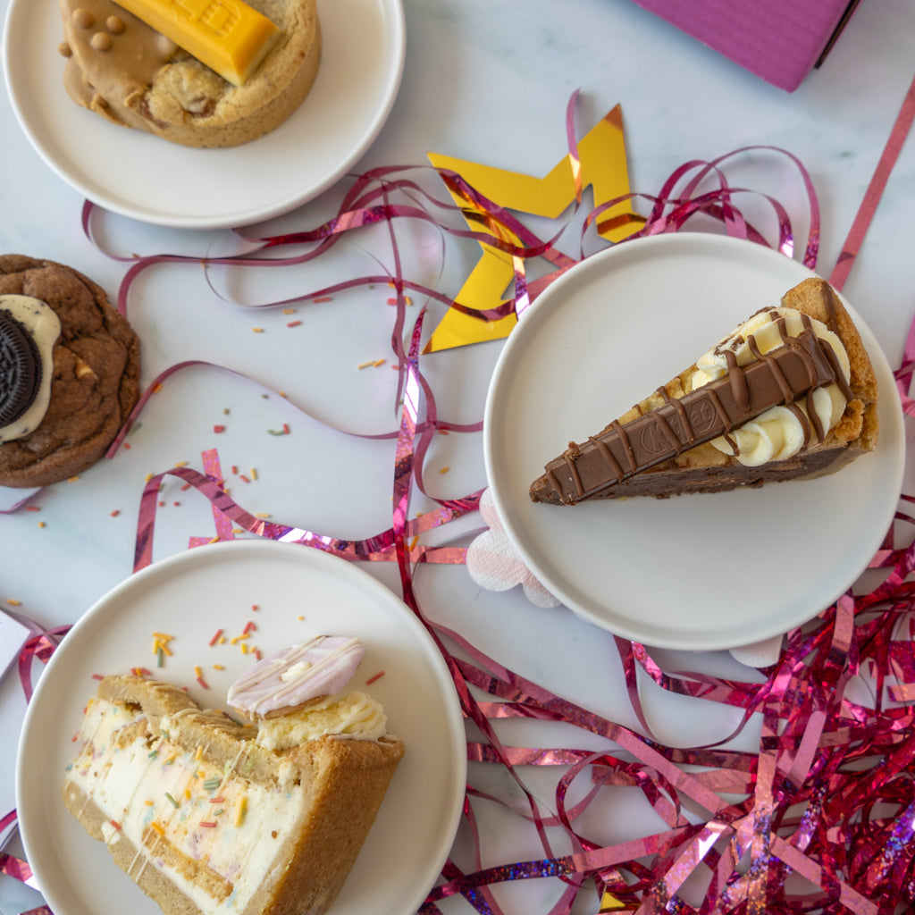 Three slices of layered cake or pastry served on white plates, arranged on a light marble-like surface with pink tinsel streamers and decorative props. The desserts include cream, chocolate, cookies, and cookie pie slices.