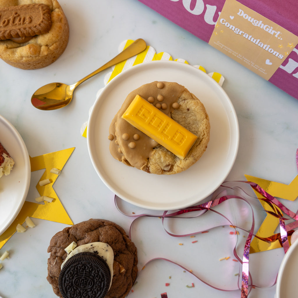 A styled flat-lay of assorted cookies on white plates with a gold spoon, yellow star confetti, and a pink box labeled DoughGirl.
