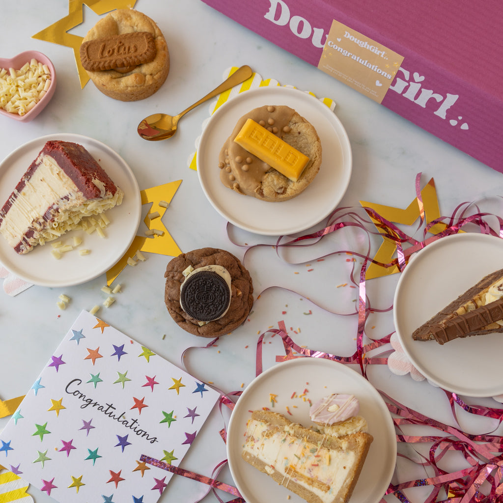 A styled flat-lay of birthday treats and a pink 'DoughGirl' gift box on a white surface, surrounded by plates of cake slices, cookies with chocolate and caramel toppings, a small bowl of shredded cheese, a gold spoon, star-shaped decorations, and a colorful congratulations card.