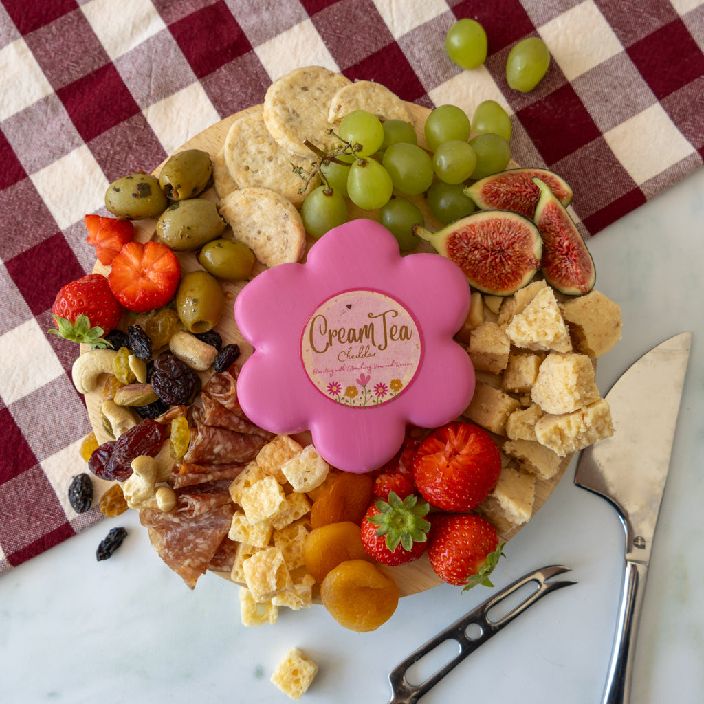 A beautifully arranged charcuterie board on a white marble surface with a red and white checkered cloth. The board is filled with crackers, green grapes, figs, strawberries, olives, cheese cubes, salami, and dried fruits, and is topped with a pink flower-shaped container labeled 'Cream Tea Cheddar'.