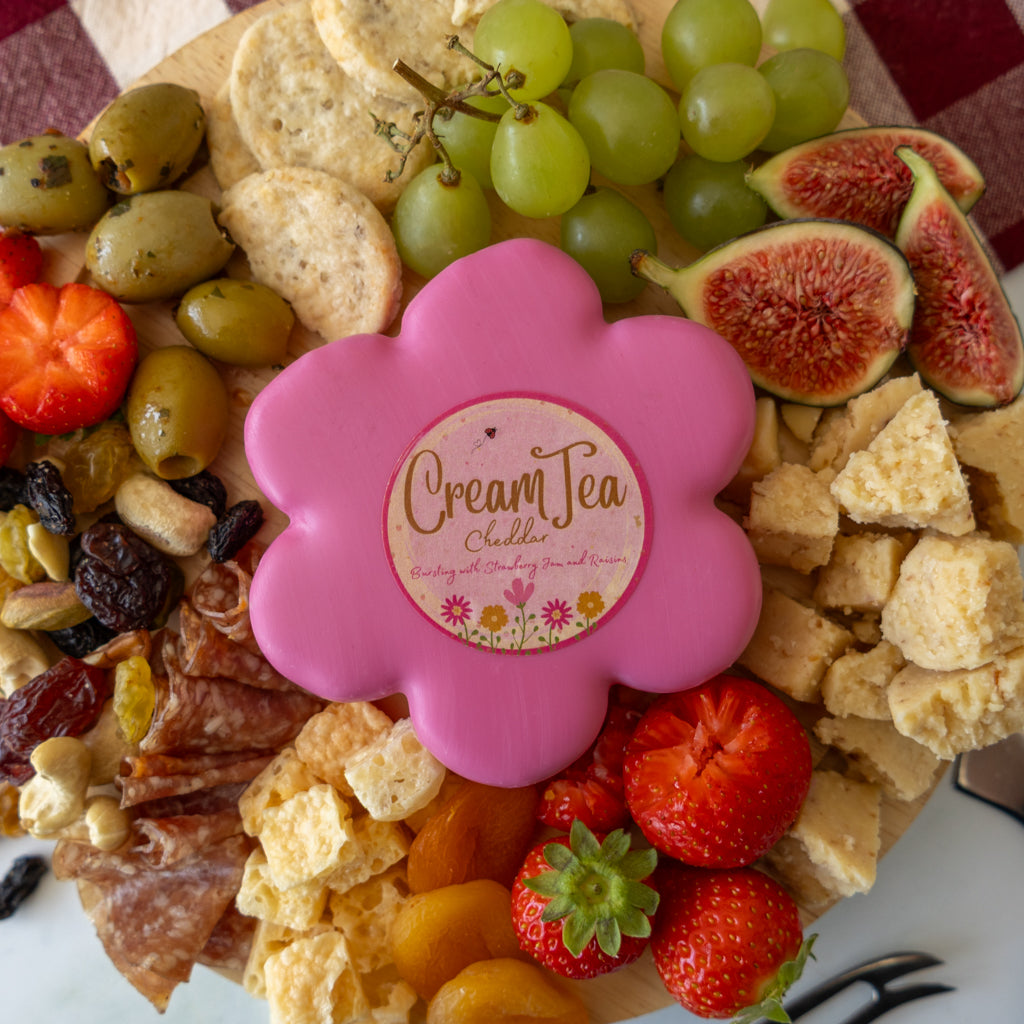 A close-up overhead shot of a charcuterie board topped with a pink flower-shaped container labeled 'Cream Tea Chilli.' The board includes grapes, figs, strawberries, olives, crackers, cheese, salami, and dried fruit on a red-and-white checkered cloth background.
