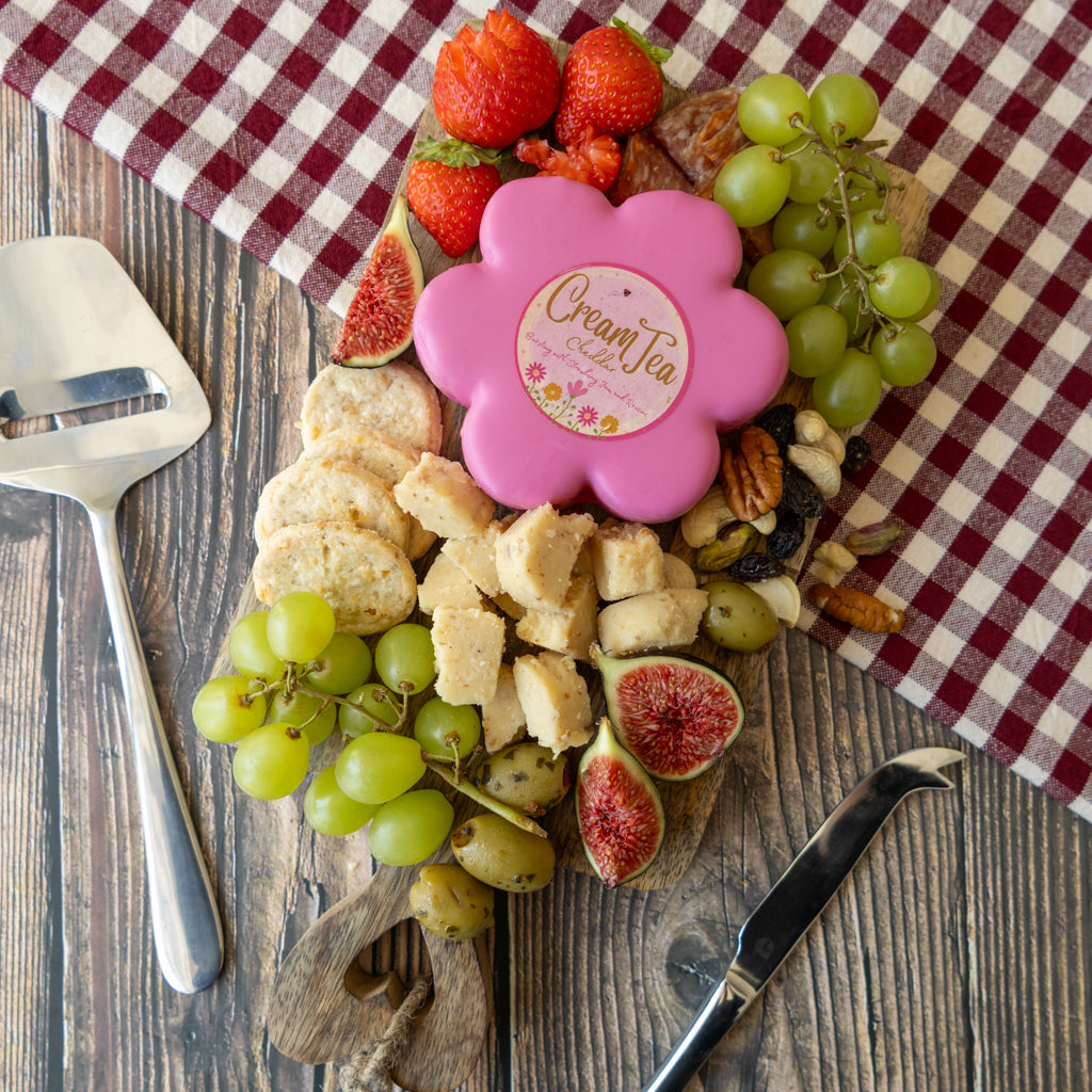 A gourmet charcuterie board styled on a rustic wooden table with a red and white checkered cloth. The board is filled with sliced cheeses, green grapes, strawberries, figs, walnuts, and dried fruits, with a pink flower-shaped soap dish in the center featuring the text 'Creamy Tea'.