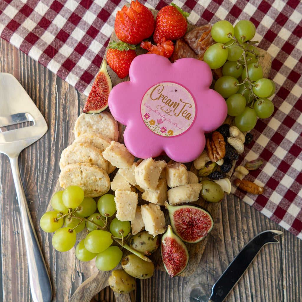 A pink flower-shaped Cream Tea gift box sits on a wooden board filled with strawberries, green grapes, figs, crackers, and nuts, with a red-and-white checkered cloth underneath.