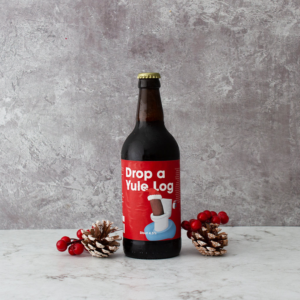 “Drop a Yule Log” Christmas beer bottle styled with pine cones and red berries.
