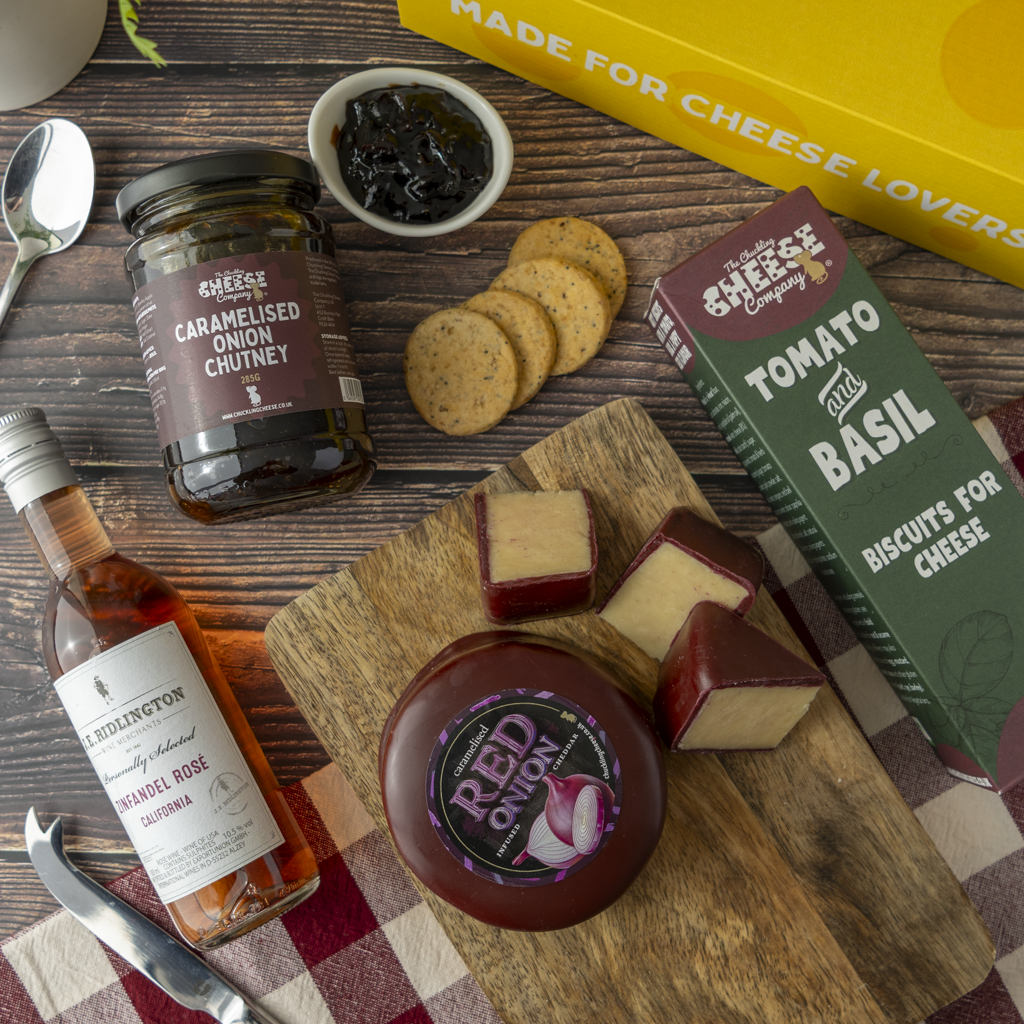 A styled cheese and biscuit gift assortment on a wooden board with crackers, a bottle of Ridlington Rosé, a jar of caramelised onion chutney, and a box of tomato and basil biscuits for cheese, arranged on a rustic wood background.