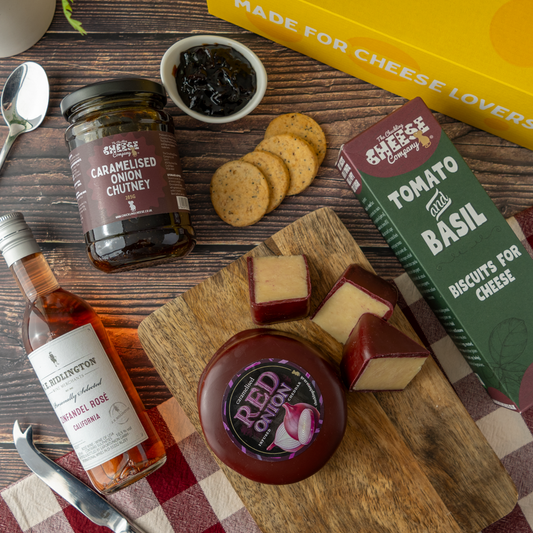 A styled cheese and biscuit gift assortment on a wooden board with crackers, a bottle of Ridlington Rosé, a jar of caramelised onion chutney, and a box of tomato and basil biscuits for cheese, arranged on a rustic wood background.