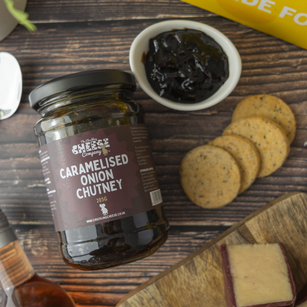 A glass jar of The Chuckling Cheese Company Caramelised Onion Chutney on a dark wooden table, styled with a small bowl of dark chutney, round crackers, and a slice of cheddar cheese.
