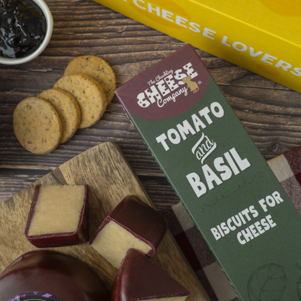 The Chuckling Cheese Company Tomato and Basil Biscuits for Cheese box shown on a wooden table with cheese wedges, round biscuits, and a yellow box in the background.