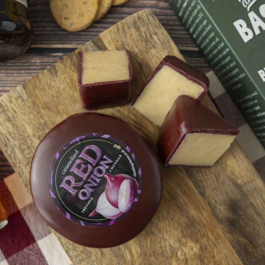A round wheel of onion cheese with a dark red-purple waxed cheese truckle and a cream-coloured interior, shown on a wooden board with two cut pieces. The cheese label reads 'Caramelised RED ONION'.