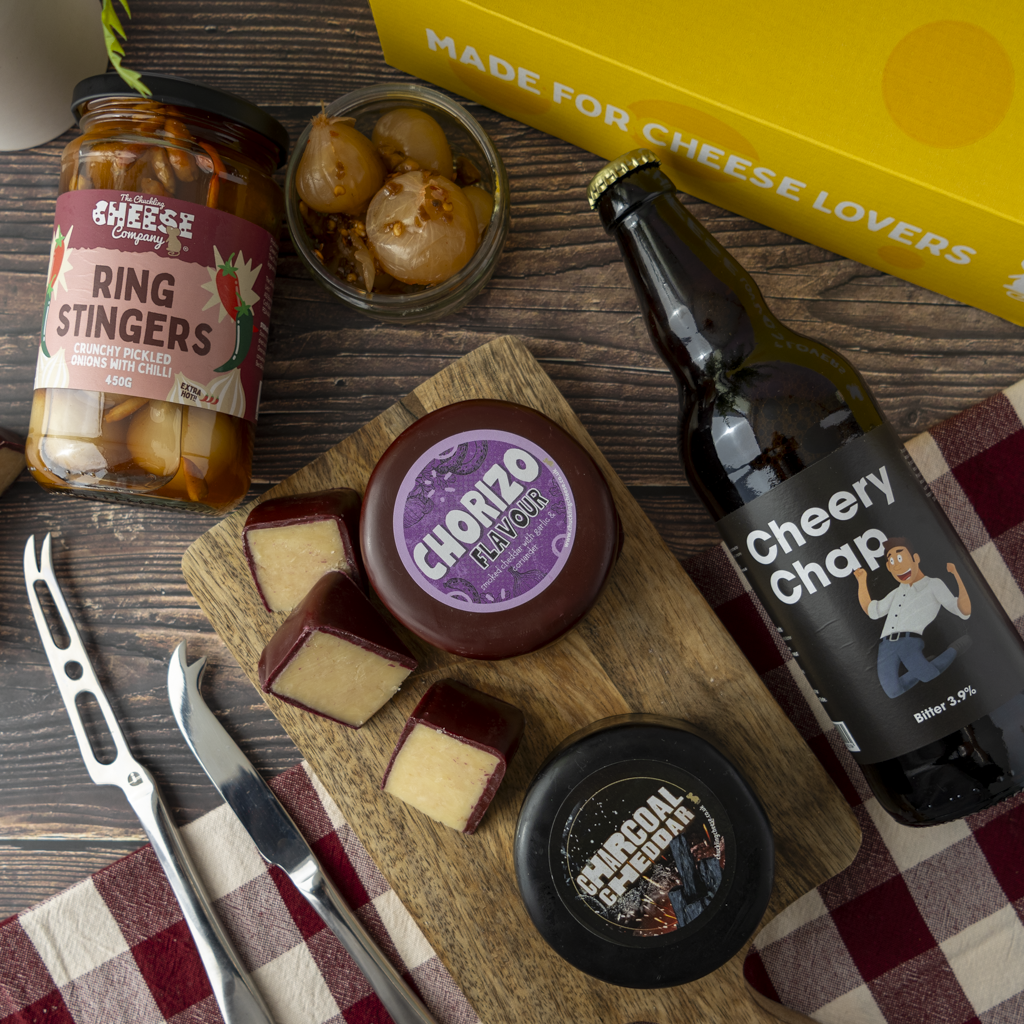 A styled flat lay of a cheese and food gift set on a wooden table, featuring a bottle of Cheery Chaps beer, a jar of The Chuckling Cheese Company Ring Stingers, a wooden board with cheese wedges and a round of Chorizo Flavour cheese, a small jar of pickled onions, cheese knives, and a yellow box printed with 'MADE FOR CHEESE LOVERS'.