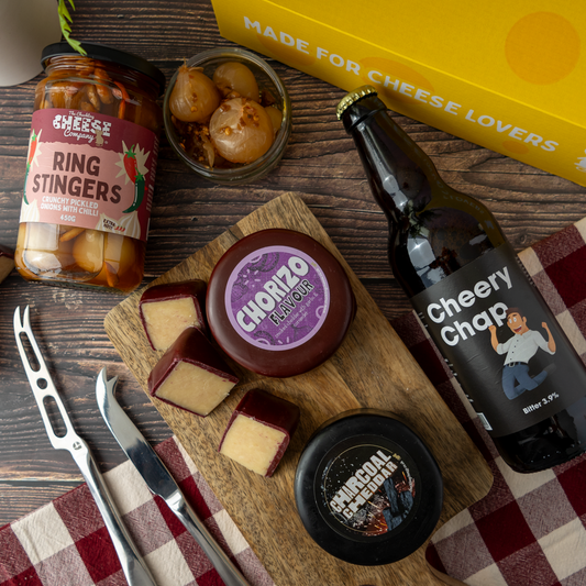 A styled flat lay of a cheese and food gift set on a wooden table, featuring a bottle of Cheery Chaps beer, a jar of The Chuckling Cheese Company Ring Stingers, a wooden board with cheese wedges and a round of Chorizo Flavour cheese, a small jar of pickled onions, cheese knives, and a yellow box printed with 'MADE FOR CHEESE LOVERS'.