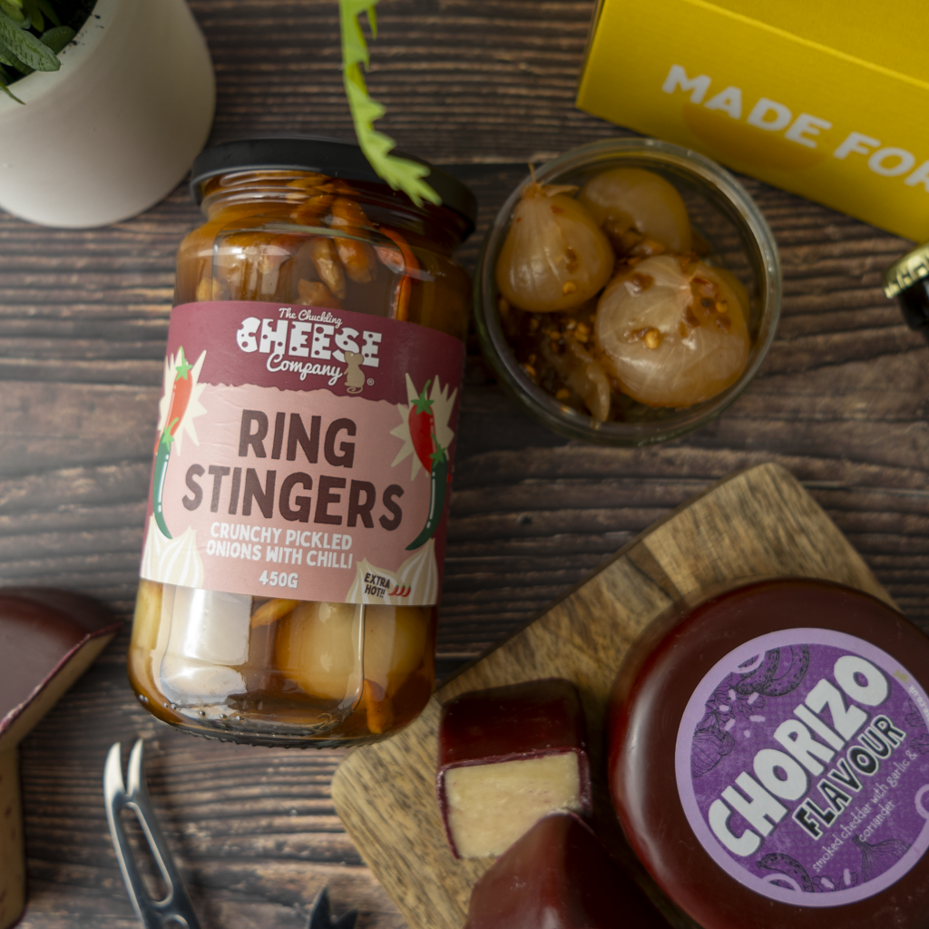 A jar of The The Chuckling Cheese Company Ring Stingers crunchy pickled onions with chilli, styled on a dark wooden table with a small bowl of pickled onions, a wheel of chorizo cheese, and a yellow box in the background.