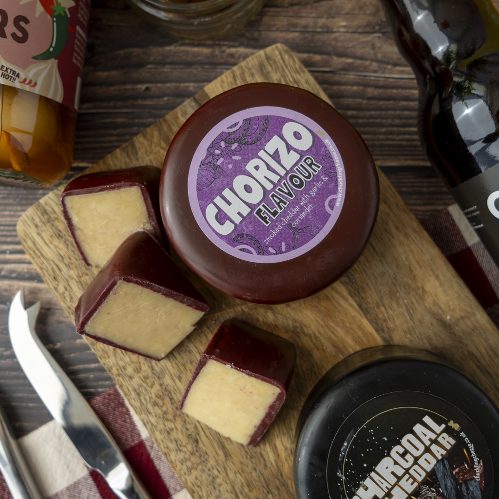 A round wheel of chorizo-flavoured cheese with several cut wedges on a wooden cutting board, alongside a cheese knife, and other food items on a dark wood table.