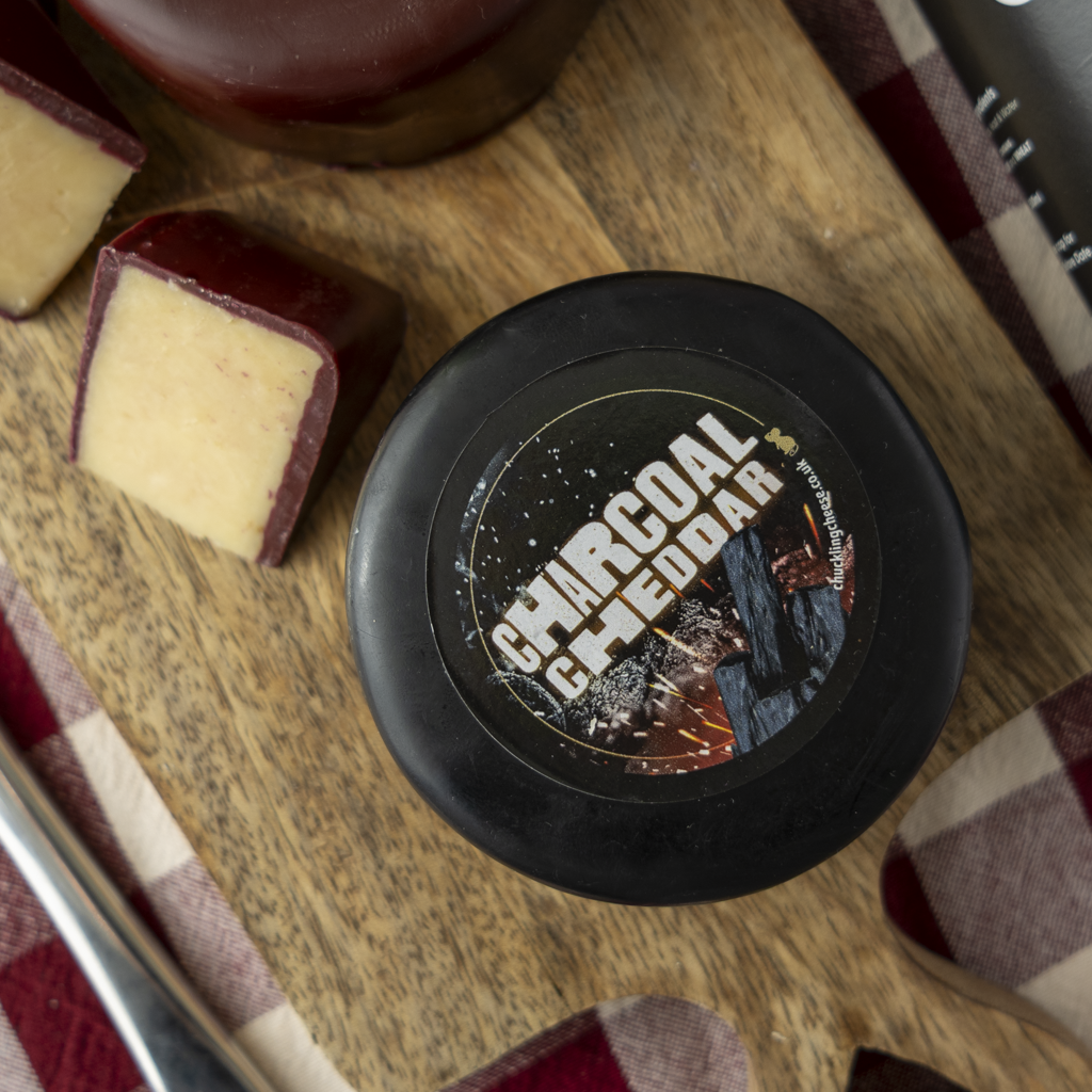 A round black container of Charcoal cheese truckle labelled 'Charcoal Cheddar' sits on a wooden board with slices of cheese and a red-and-white plaid cloth nearby.