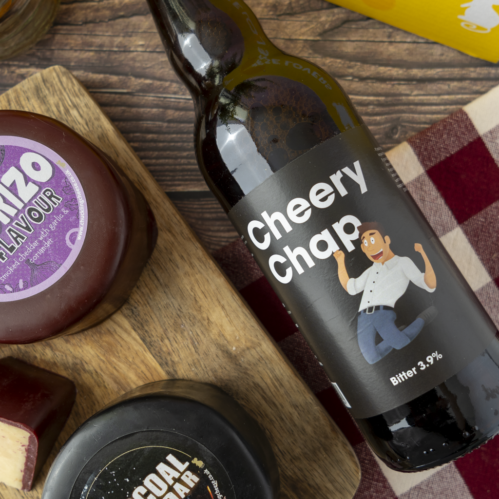 A dark glass beer bottle labeled 'Cheery Chap' with a cartoon man graphic, placed diagonally on a wooden board with cheese and a plaid cloth in a styled flat-lay setting.