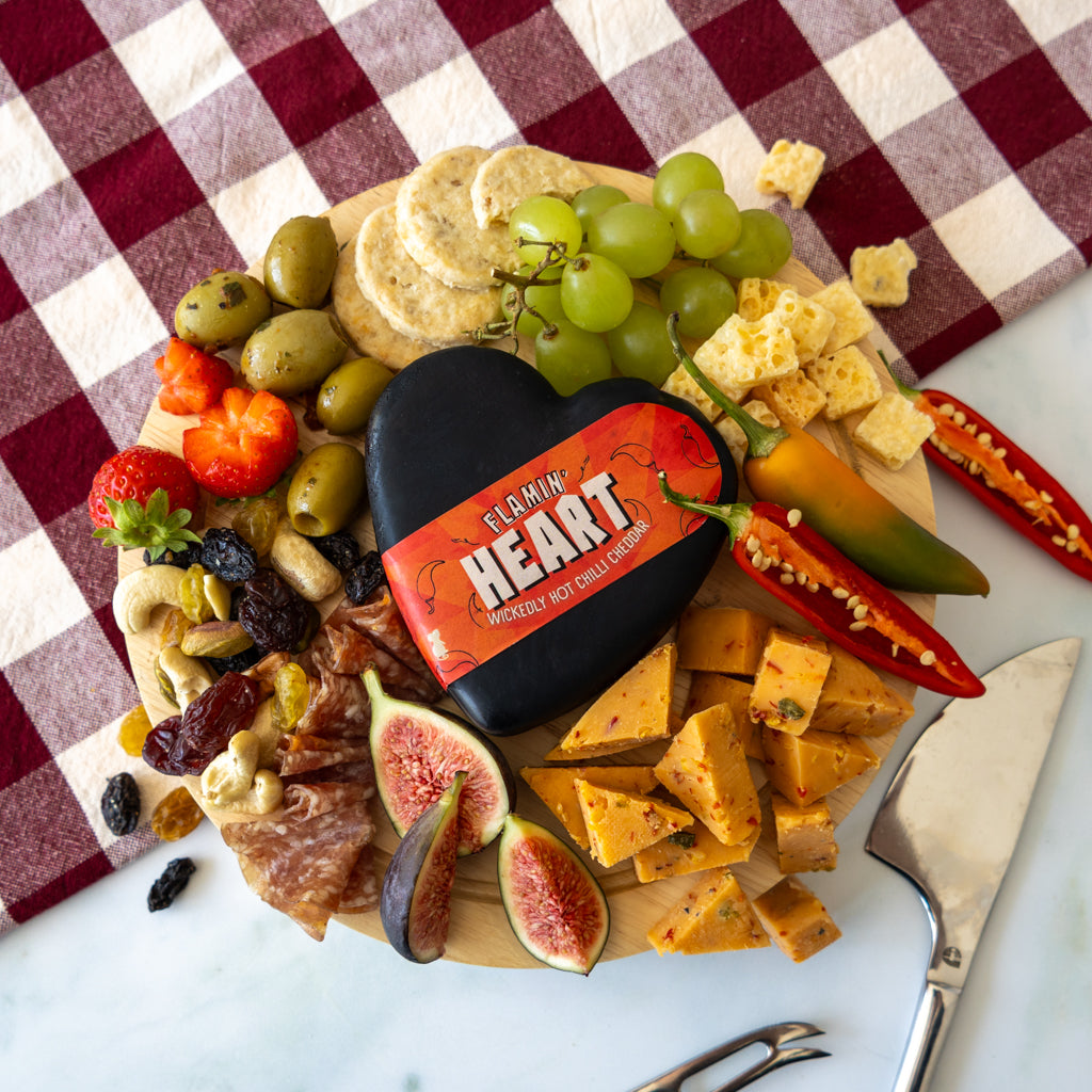 A heart-shaped charcuterie board filled with cheese, grapes, olives, crackers, fruit, and a black heart-shaped cheese block labeled 'FLAMMIN HEART'. The board is styled on a red-and-white checkered cloth over a white marble surface with a knife nearby.