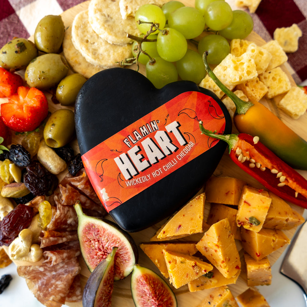 A heart-shaped black cheese wheel with a bright orange-red label reading 'FLAMIN' HEART' is arranged on a charcuterie board with grapes, olives, crackers, peppers, figs, and cured meats.