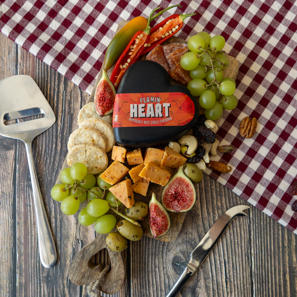 A charcuterie board arrangement on a wooden surface with a red-and-white gingham cloth. The centerpiece is a black cheese block labeled 'FLAMMIN HEART', surrounded by green grapes, sliced figs, crackers, nuts, and peppers.