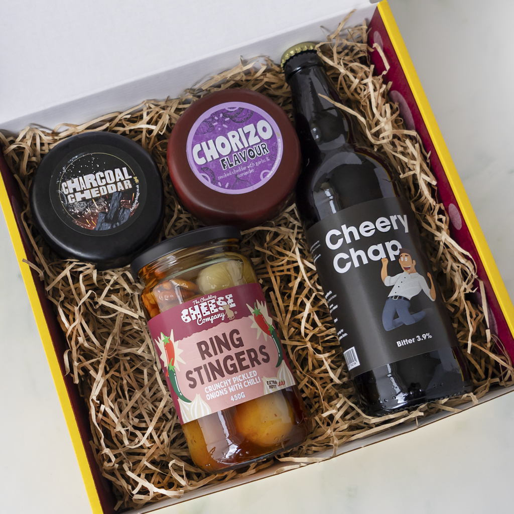 A curated gift box filled with snacks and treats, including a bottle of Cheery Chap beer, a jar of Ring Stingers, a round cheese labeled Chorizo Flavour, and a black container labeled Charcoal & Spiced.
