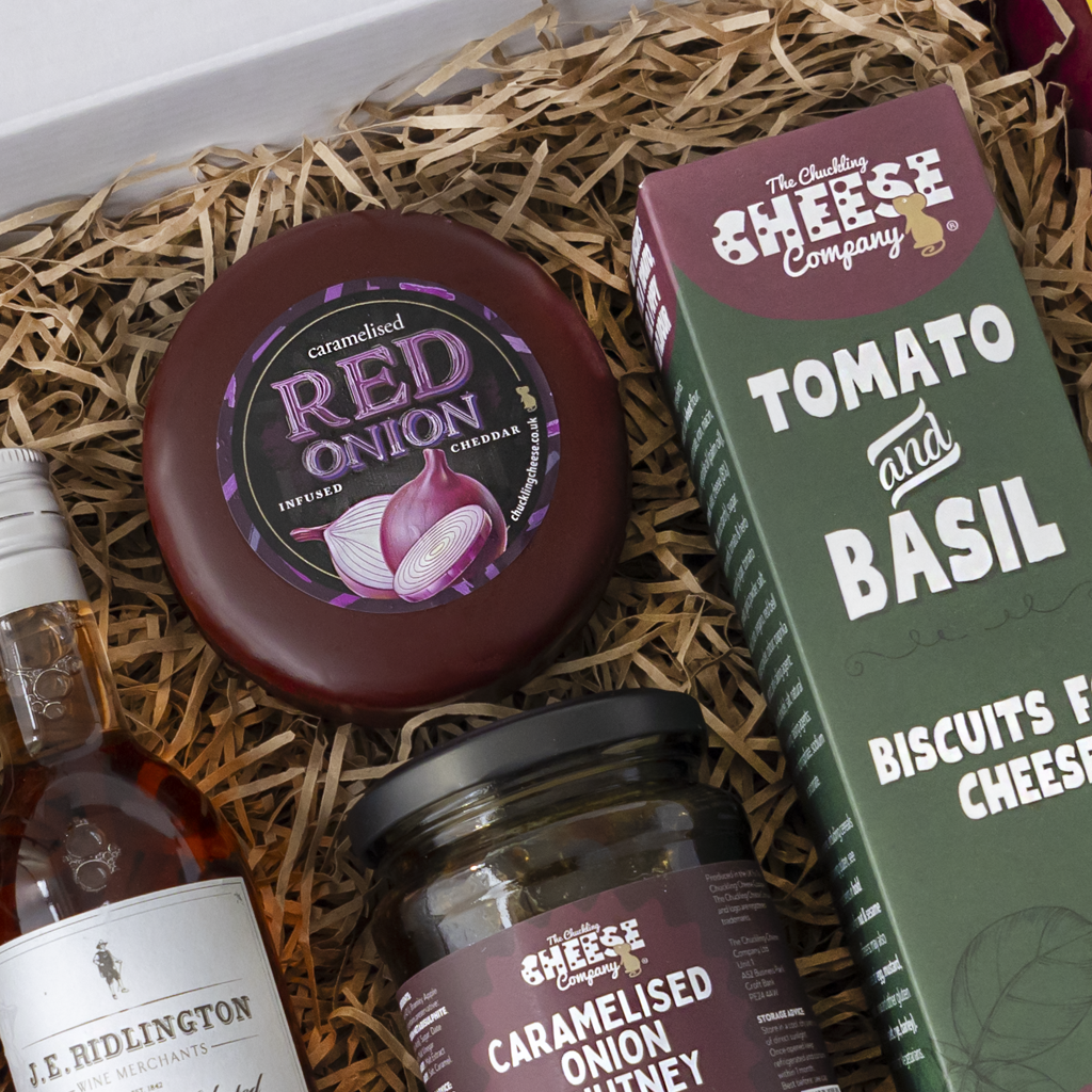 A curated cheese and snack gift box from The Chuckling Cheese Company, featuring a round caramelised red onion cheese wheel, a jar of caramelised onion chive, a box of tomato and basil biscuits, and a bottle of J.B.