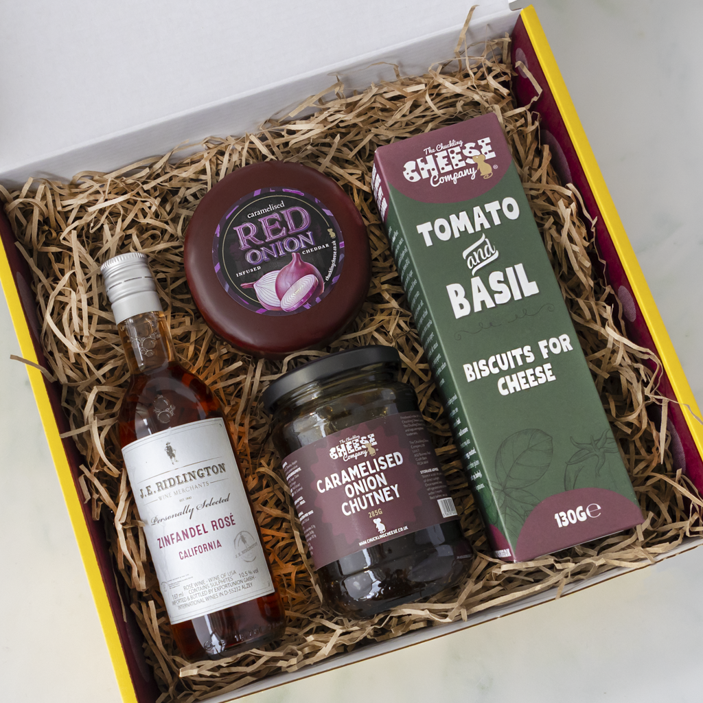 A curated cheese and food gift box filled with shredded paper, containing a bottle of J.B., a jar of chutney, a cheese truckle, and a tomato and basil biscuit for cheese box