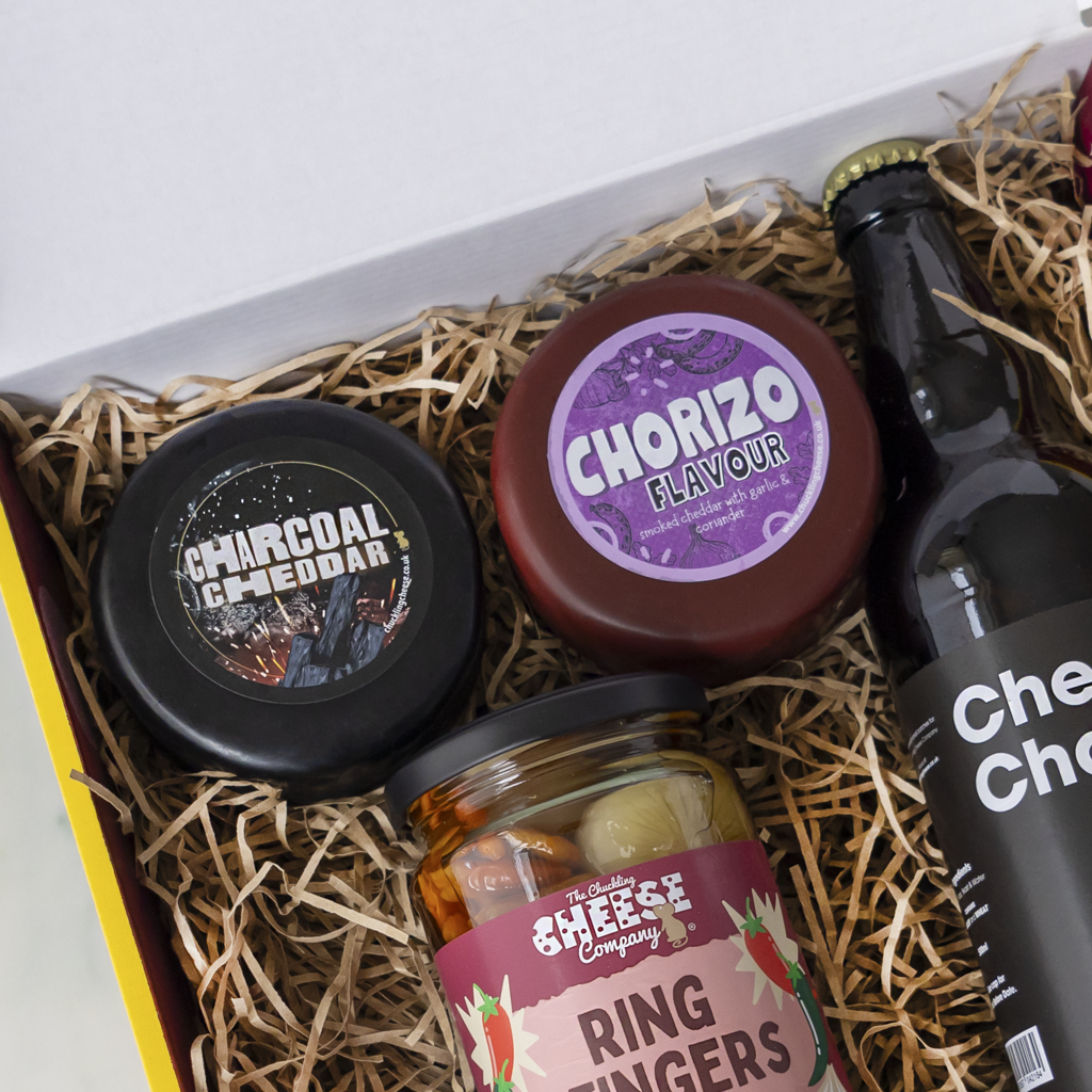 A curated food gift box filled with shredded straw, containing a jar of The Chuckling Cheese Company ring fingers, a round jar of charcoal and cheddar, a round tin of chorizo flavour, and a bottle of cheese and chocolate.