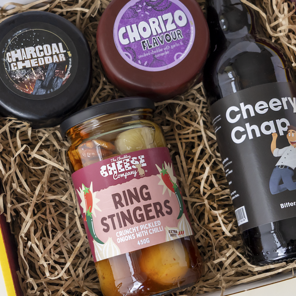 A curated food gift box filled with shredded paper, featuring a jar of The Chuckling Cheese Company Ring Stingers pickled onions with chilli, a round package of Chorizo Flavour cheese, a tin of Charcoal Cheddar, and a bottle of Cheery Chap beer.