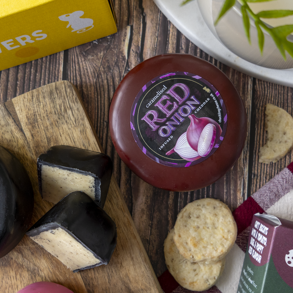 A round truckle of red onion-flavoured cheese with a dark red-brown outer rind and a black label reading 'RED ONION', styled on a wooden surface with sliced cheese, crackers, and partial packaging visible.