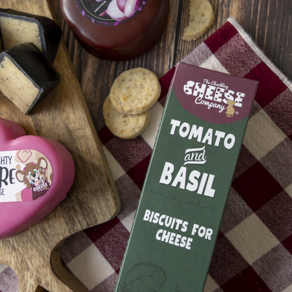 A boxed food product labelled 'The Chuckling Cheese Company Tomato and Basil Biscuits for Cheese' placed on a red-and-white checkered cloth over a wooden surface, with cheese wedges, crackers, and a bottle nearby.