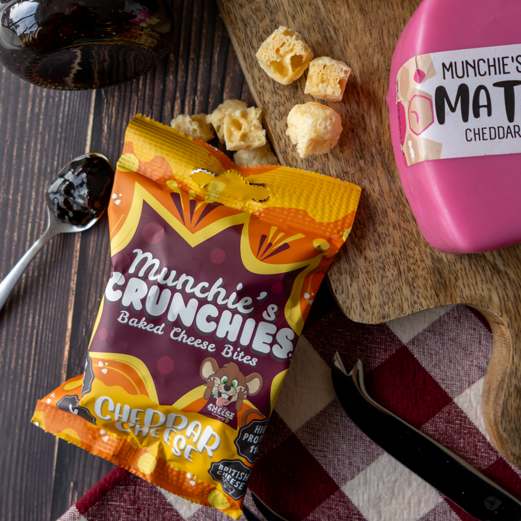 A bag of Munchie's Crunchies Baked Cheese Bites in Cheddar flavor, styled on a dark wood surface with a pink lunchbox, crackers, a spoon with cheddar sauce, and a checkered cloth.