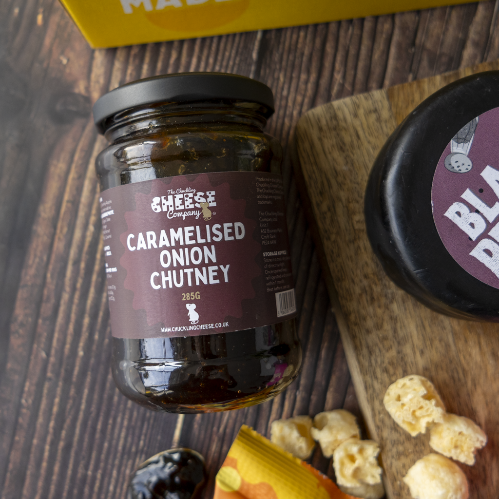 A jar of The Chuckling Cheese Company Caramelised Onion Chutney on a dark wooden table, styled with a wheel of black cheese, dried onion pieces, and a yellow box in the background.