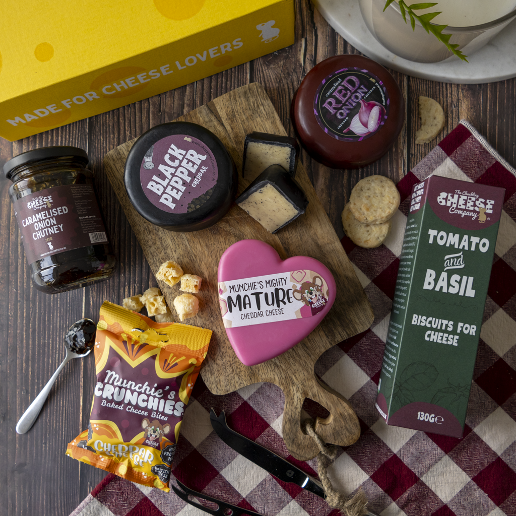 A styled cheese and snack gift arrangement on a dark wood surface, featuring multiple cheese products, crackers, a bag of Munchie's Crunchy, a yellow box, and a red-and-white check cloth.