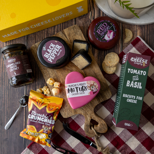A styled cheese and snack gift arrangement on a dark wood surface, featuring multiple cheese products, crackers, a bag of Munchie's Crunchy, a yellow box, and a red-and-white check cloth.