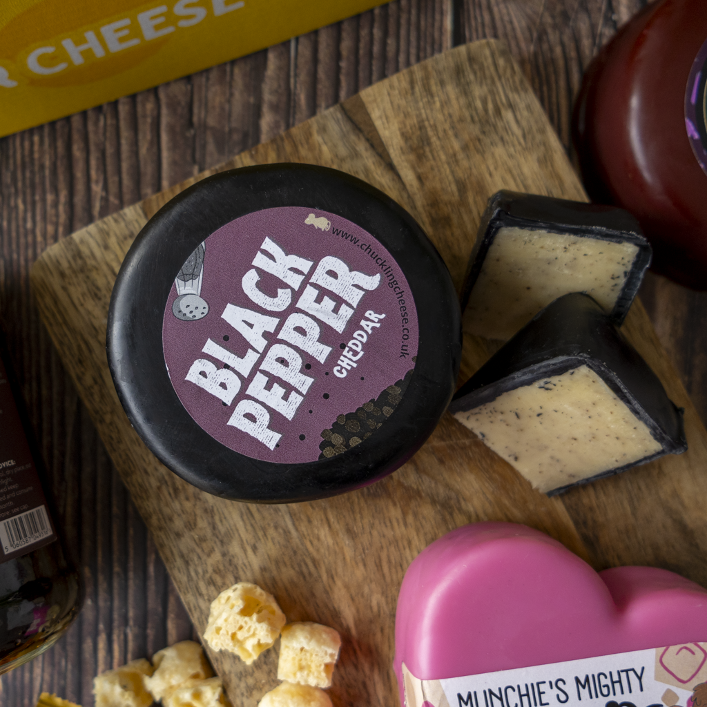 A round wheel of Black Pepper Cheddar cheese with its dark rind and purple label, displayed on a wooden cutting board with two wedges cut and showing the pale cheddar interior. Surrounding props include a pink heart-shaped container, cheese cubes, and other food items on a dark wood surface.