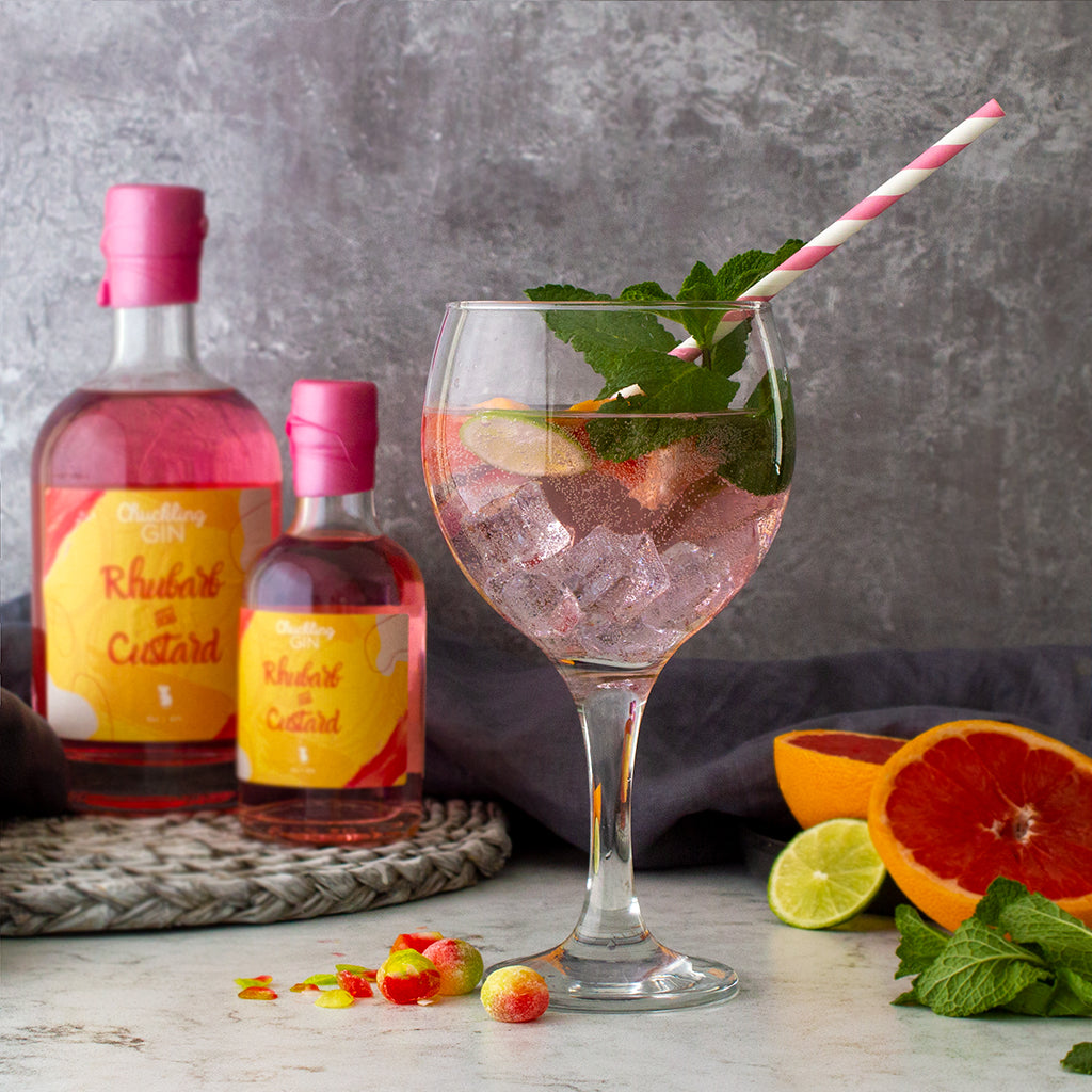 A glass of pink rhubarb and custard cocktail with ice, mint leaves, and a pink-and-white striped straw, styled with two bottles of rhubarb and citrus slices on a grey surface against a textured grey background.