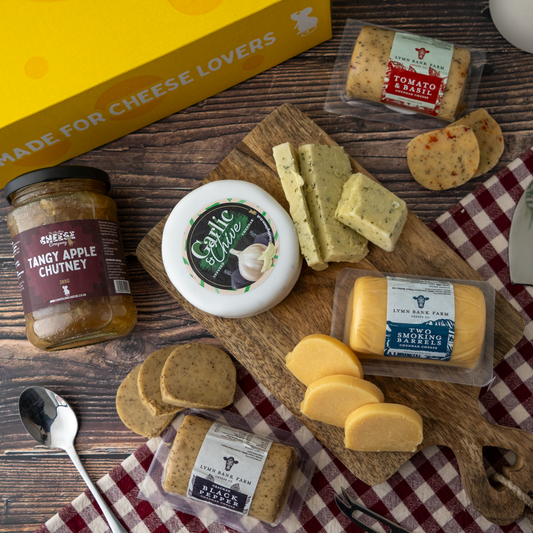 A cheese and charcuterie gift set arranged on a wooden surface, including a jar of tangy apple chutney, a round of garlic cheese, several sliced cheeses, crackers, and a yellow box labeled 'Made for Cheese Lovers'.