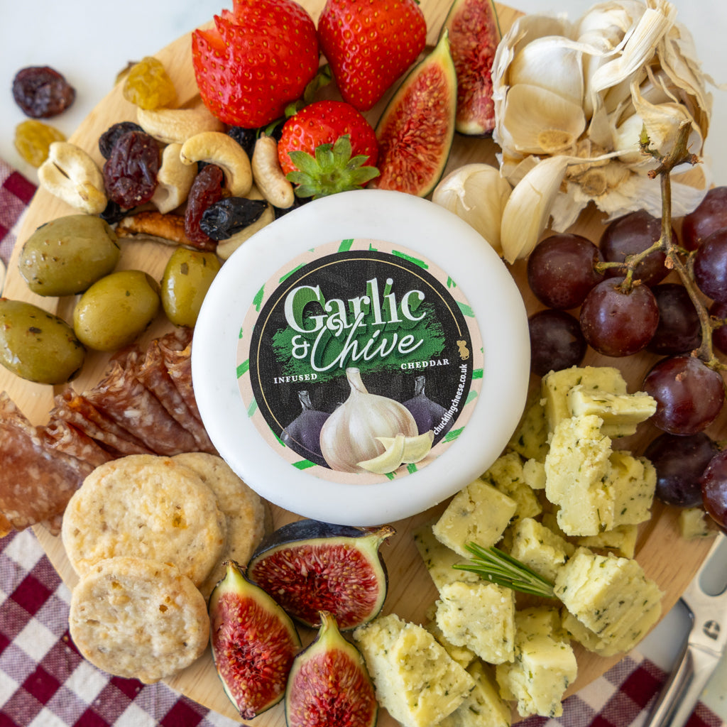 A round white container of Garlic & Chive Infused Cheese sits on a wooden charcuterie board surrounded by sliced figs, strawberries, grapes, olives, crackers, salami, garlic cloves, and cheese cubes on a red-and-white checkered cloth.