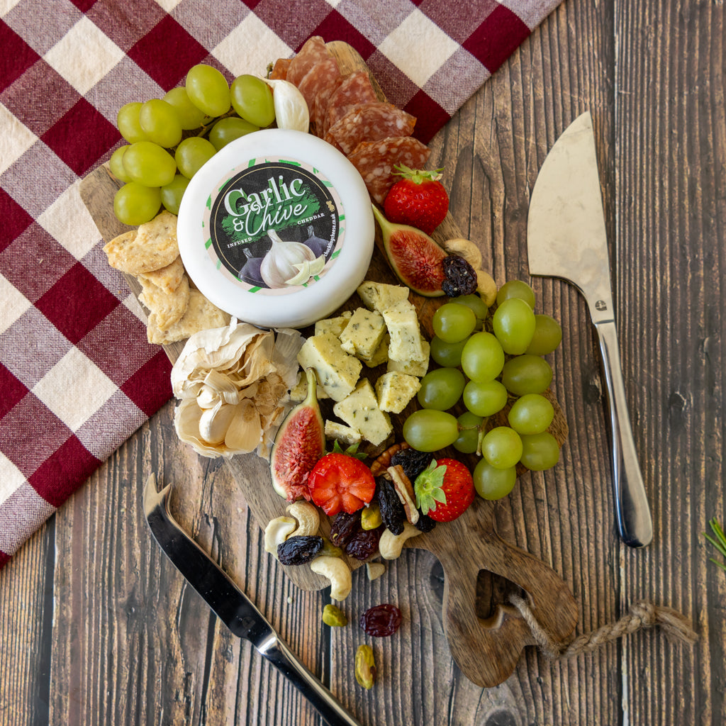 A charcuterie board arrangement on a wooden surface featuring a round container of Garlic Chive cheese, green grapes, sliced strawberries, figs, crackers, nuts, dried fruit, salami, and two knives, with a red-and-beige checkered cloth in the background.