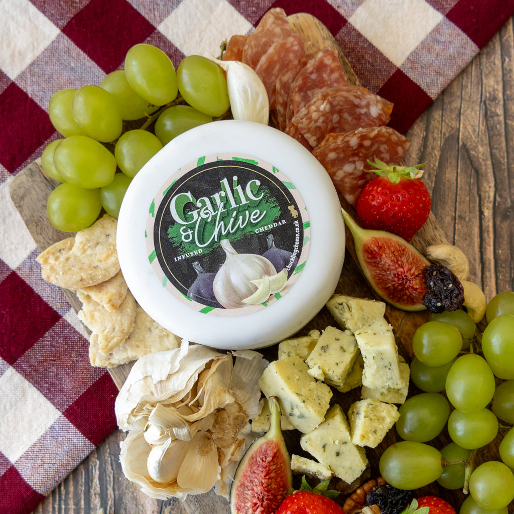A round white cheese wheel labeled 'Garlic & Chive' surrounded by an appetizing charcuterie board arrangement on a red-and-white checkered cloth over a wooden surface, including green grapes, sliced salami, crackers, blue cheese, garlic, figs, strawberries, and nuts.