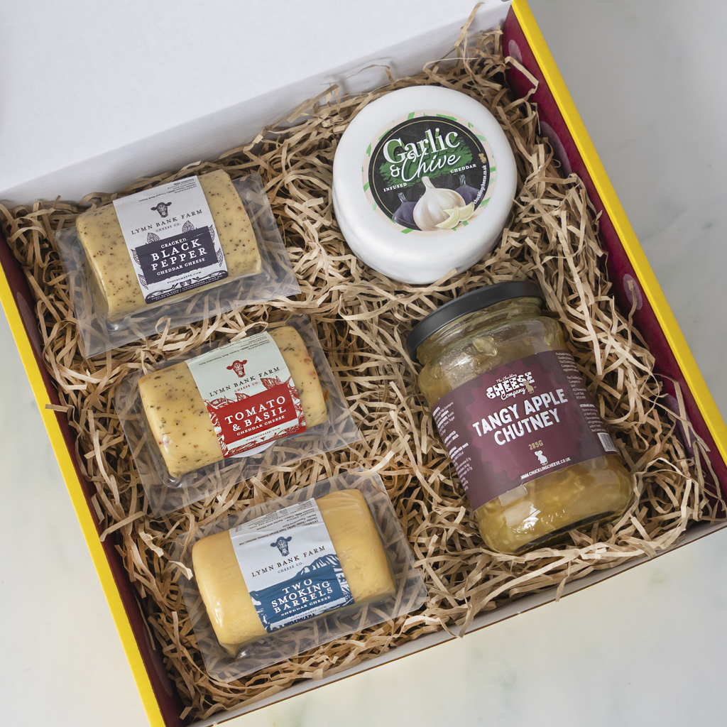 A curated cheese and pickle gift box from The Chuckling Cheese Company, filled with shredded paper straws. The box contains a round wheel of Garlic Chive cheese, a jar of Tangy Apple Chutney, and three individually wrapped blocks of cheese: Black Pepper, Tomato & Basil, and Two Smoking Cheese.
