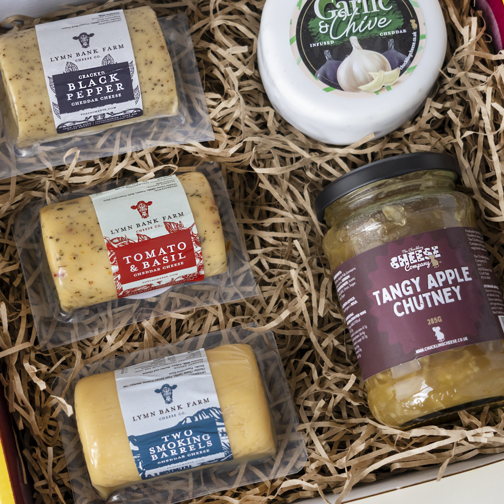 A curated cheese and charcuterie gift box from Lymn Bank Farm, featuring three packaged cheeses, a jar of tangy apple chutney, and a round container of Garlic & Chive spreads, arranged in a box filled with shredded paper.