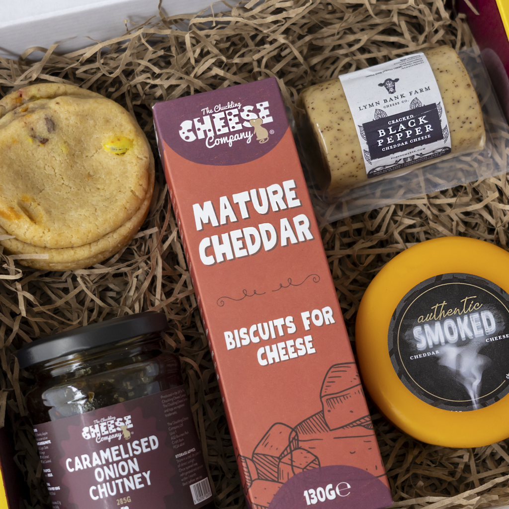 A gourmet cheese and biscuit gift box filled with shredded straw. Visible items include a tall box of The Chuckling Cheese Company Mature Cheddar Biscuits for Cheese, a round wheel of authentic smoked cheese, a jar of caramelised onion chutney, and a packaged stick of Lynn Barn Farm Black Pepper Biscuits.