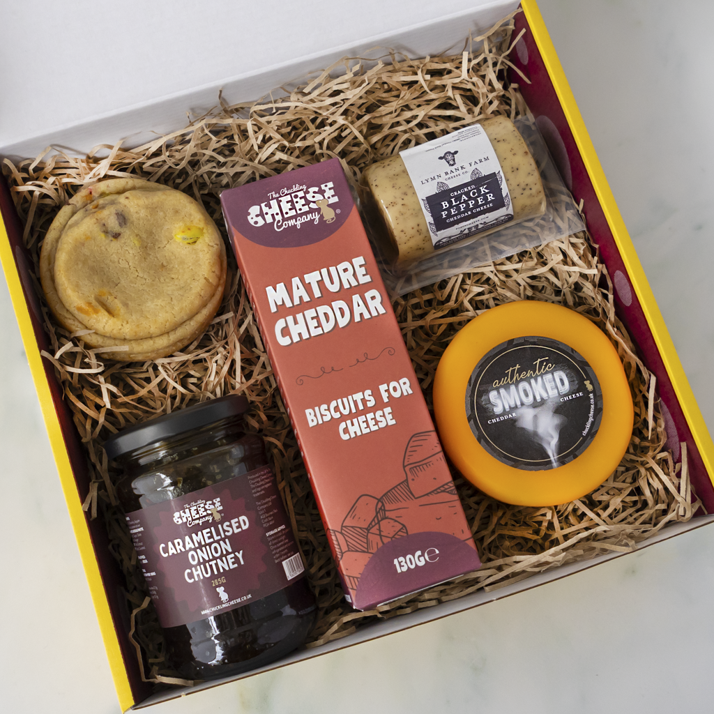 A curated cheese and biscuit gift box from The Chuckling Cheese Company, shown from a top-down view inside its yellow and white packaging. The box is filled with shredded paper and contains a round cookie, a jar of caramelised onion chutney, a box of mature cheddar cheese, a wrapped black pepper barrel, and a round wheel of smoked cheese.