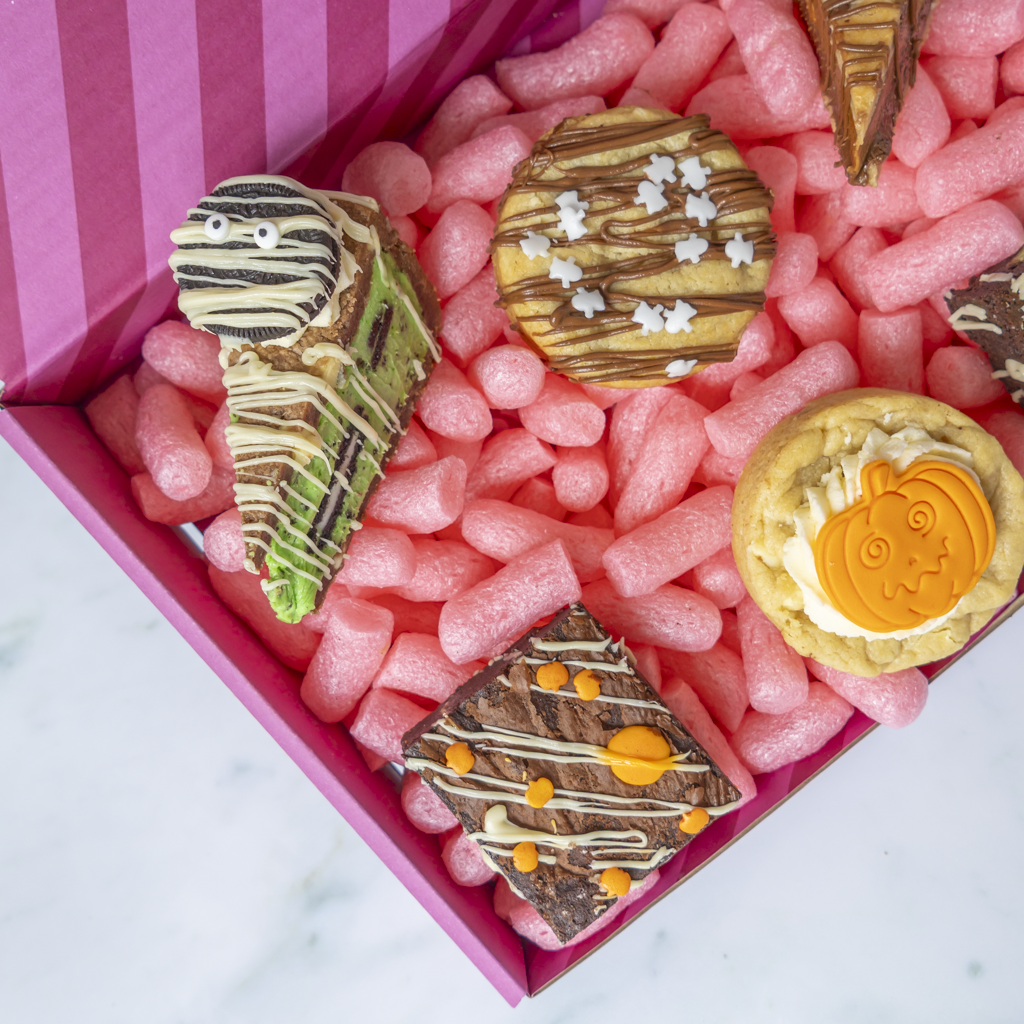 A close-up overhead view of a pink gift box filled with pink packing peanuts and decorated Halloween-themed treats, including a mummy cookie pie slice, a pumpkin cookie, and chocolate-dripped square brownies.