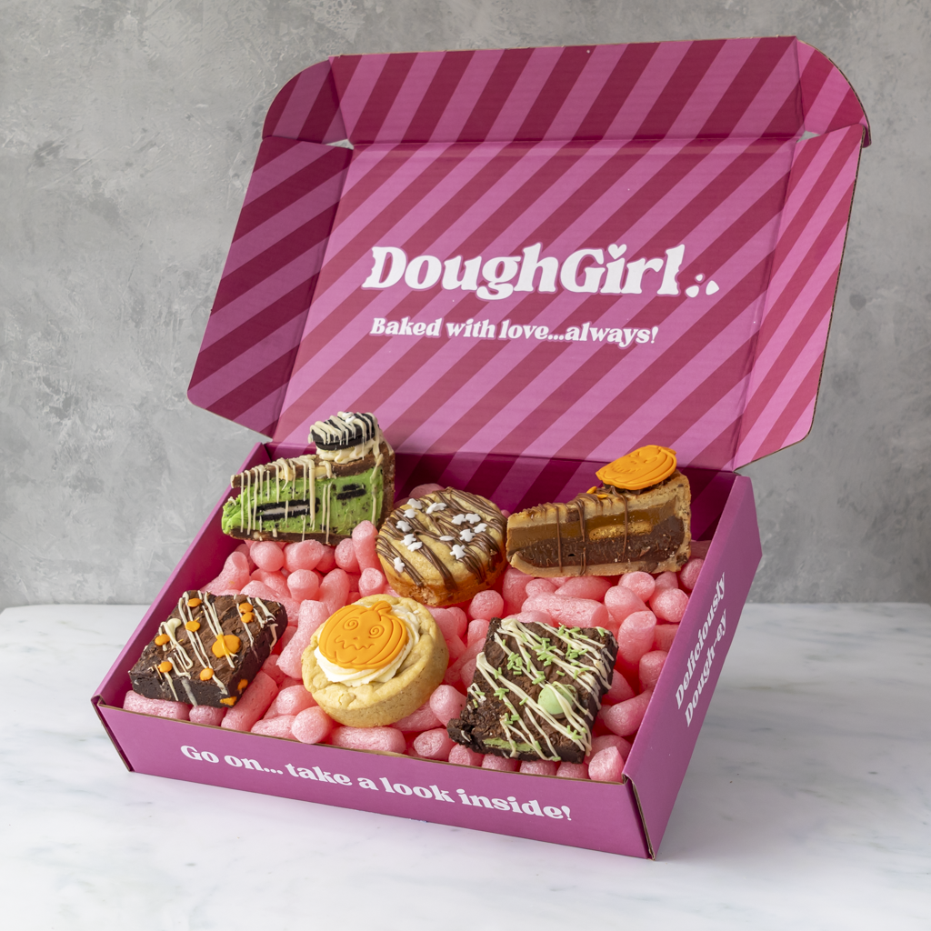 An open pink DoughGirl gift box filled with assorted gourmet pastries and cookies, set on a light marble surface against a gray wall. The box lid displays the DoughGirl brand name and slogan.