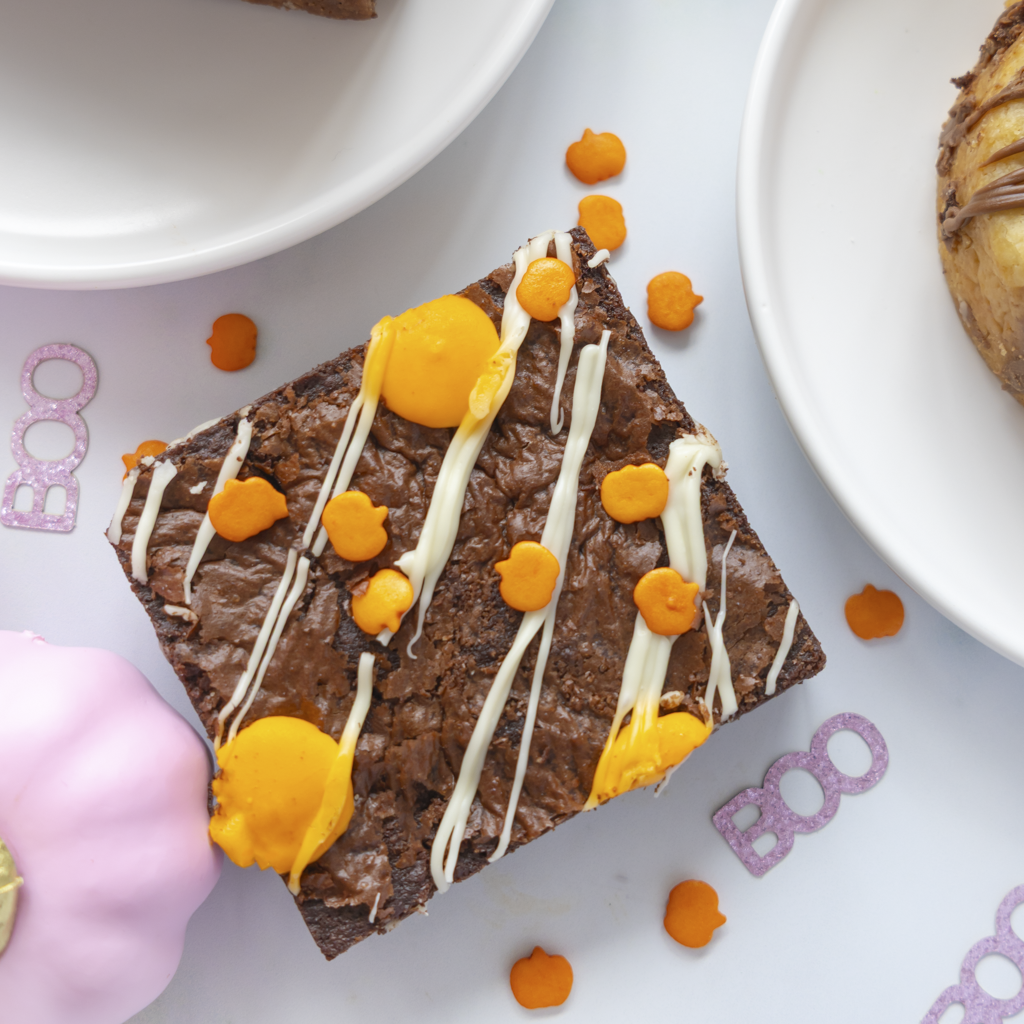 A square chocolate brownie topped with white chocolate drizzle, bright orange candy pieces, and two large orange candy drops, photographed on a white surface with partial plates and decorative pink accents around it.