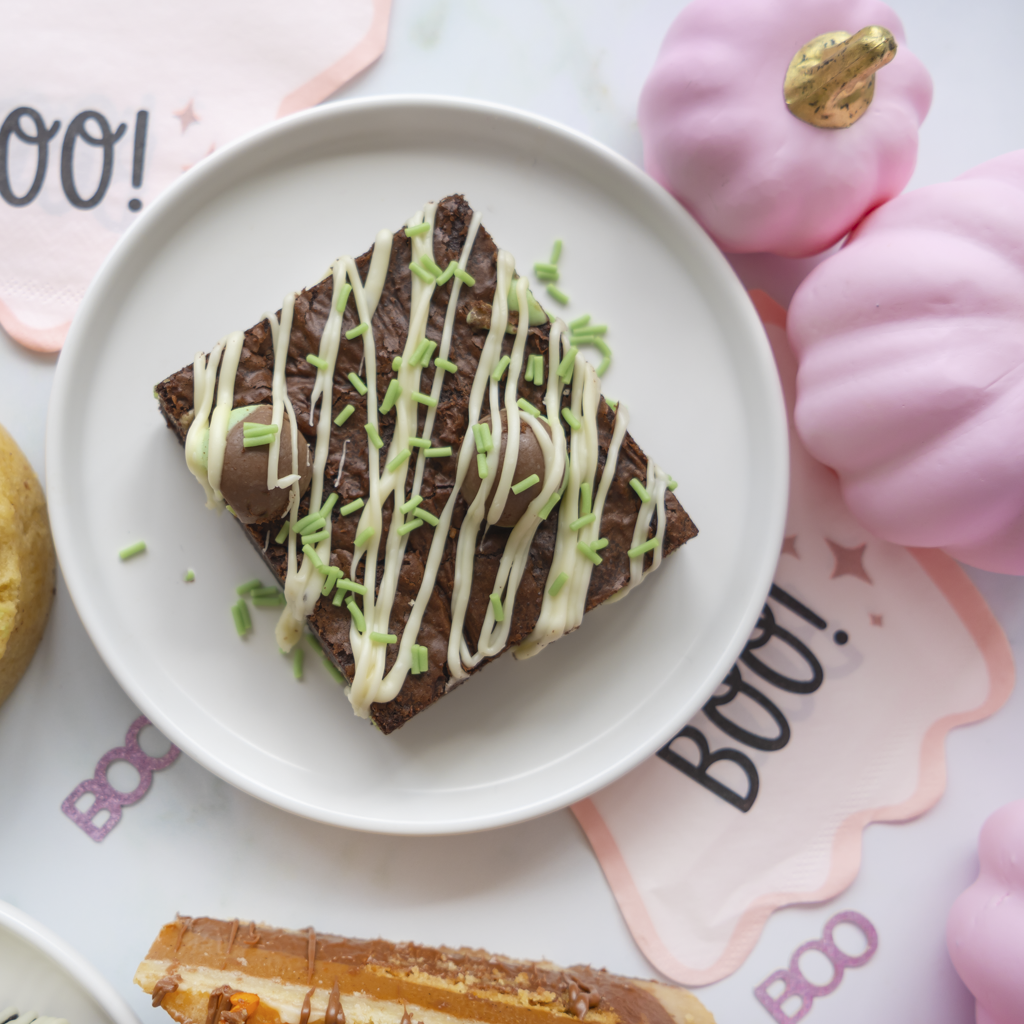 A square brownie on a white plate, decorated with white chocolate drizzle, green sprinkles, and a pumpkin spice filling, surrounded by pink decorative props and fall-themed cards.